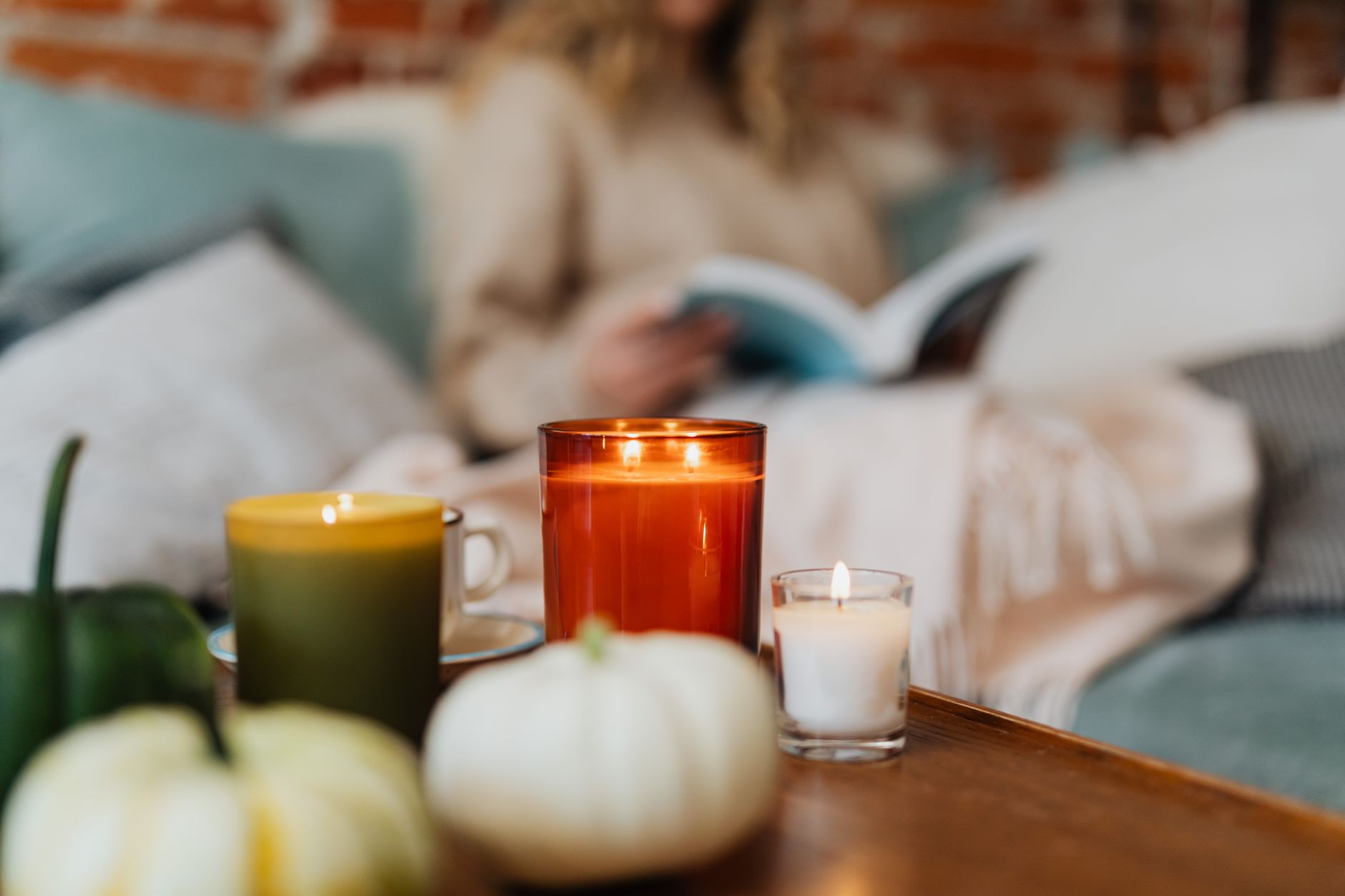 Lit candles and white pumpkins on a coffee table, someone reading on the sofa under a throw